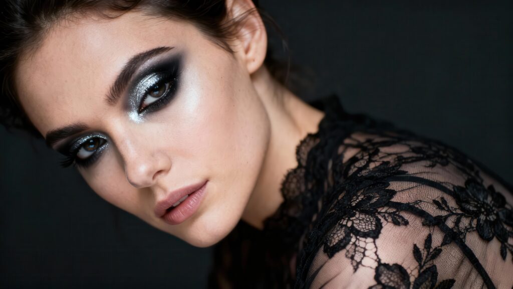 Photo of a gorgeous woman featuring heavy, dramatic black and silver glitter eyeshadow and subtle pink lip color, complemented by delicate floral black lace on her shoulder.