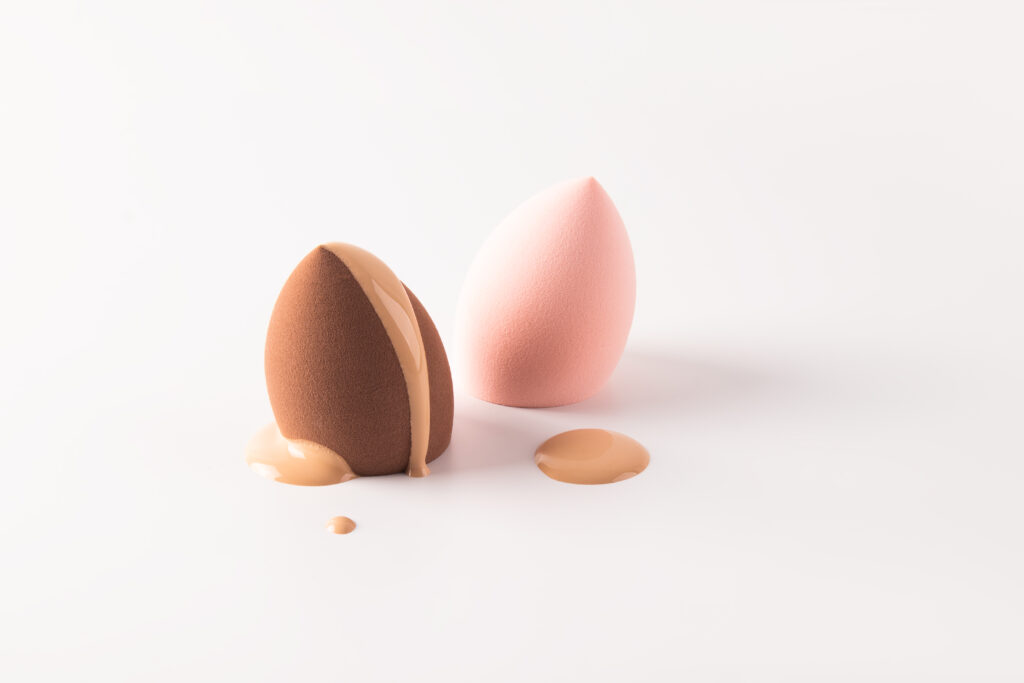 The delicate liquid texture of foundation powder in a natural tone flows down on a blender sponge. Two sponges of different colors. white background.