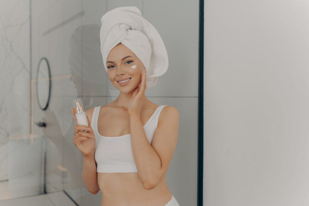 Positive young woman with head wrapped in towel smiling while applying facial cream, attractive lady using cosmetic product while doing beauty morning routine in bathroom at home. Skin care concept